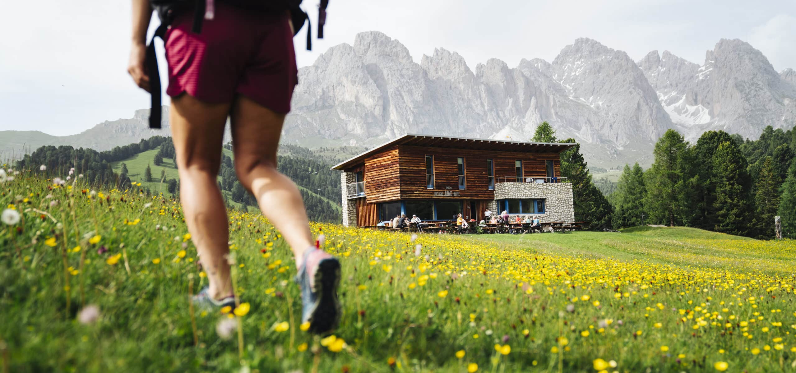 Persona con zaino cammina su prato fiorito verso rifugio in legno con persone e montagne sullo sfondo