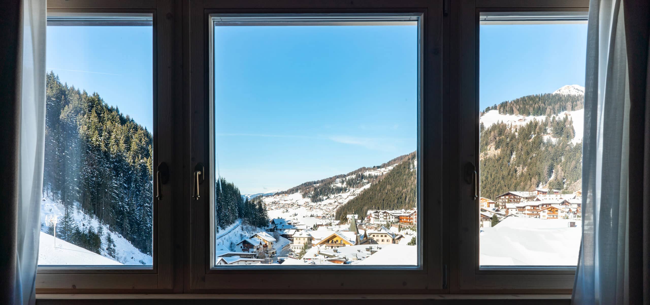 Paesino innevato con chiesa e case, tra montagne e boschi, visto da finestra a tre ante