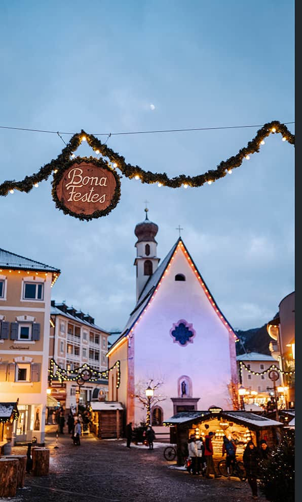 Bona Festes su ghirlanda luminosa sopra piazza con chiesa illuminata e bancarelle natalizie