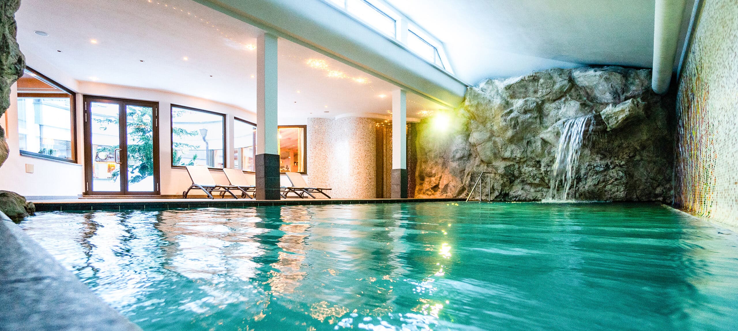 Piscina interna con cascata su parete rocciosa a destra, sdraio e grandi finestre a sinistra