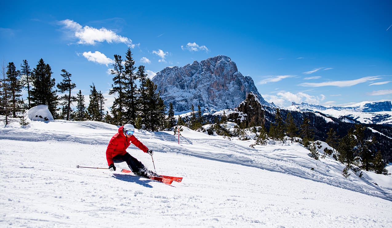 Sciatore con giacca rossa curva su pista innevata, alberi sullo sfondo e grande montagna rocciosa sotto cielo azzurro