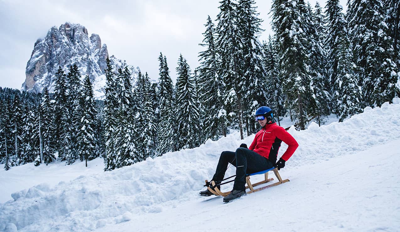 Persona con casco su slitta scende su pista innevata davanti ad abeti e una parete rocciosa