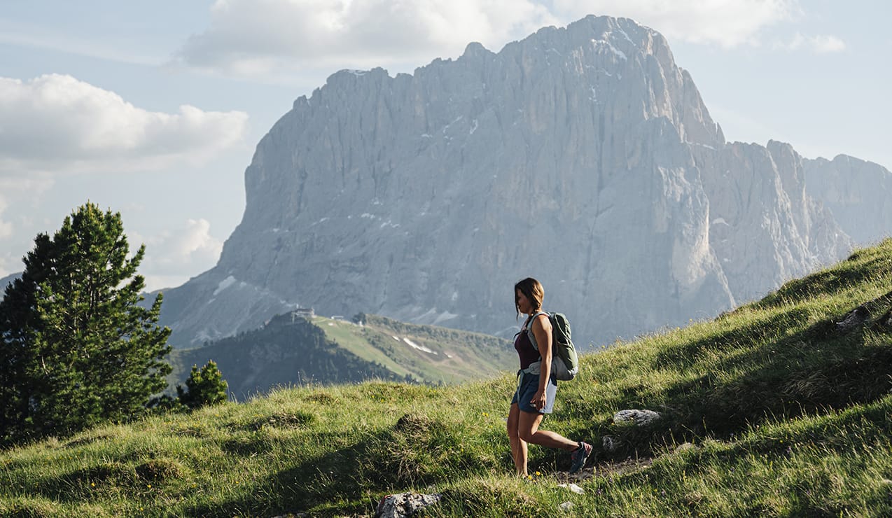 Donna con zaino che cammina su prato in montagna, grande parete rocciosa sullo sfondo.