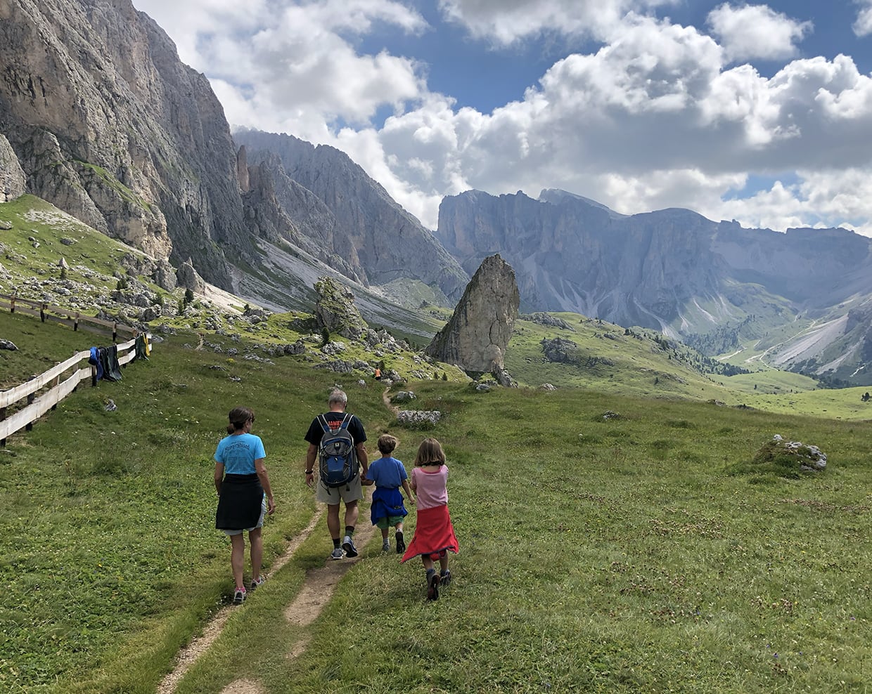 Quattro escursionisti camminano su sentiero in prato verso un grande masso verticale e montagne rocciose sullo sfondo