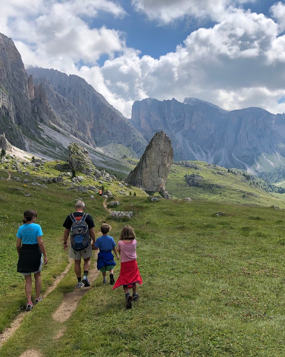 Quattro persone (due adulti, due bambini) camminano su sentiero in pascolo montano davanti a pareti rocciose