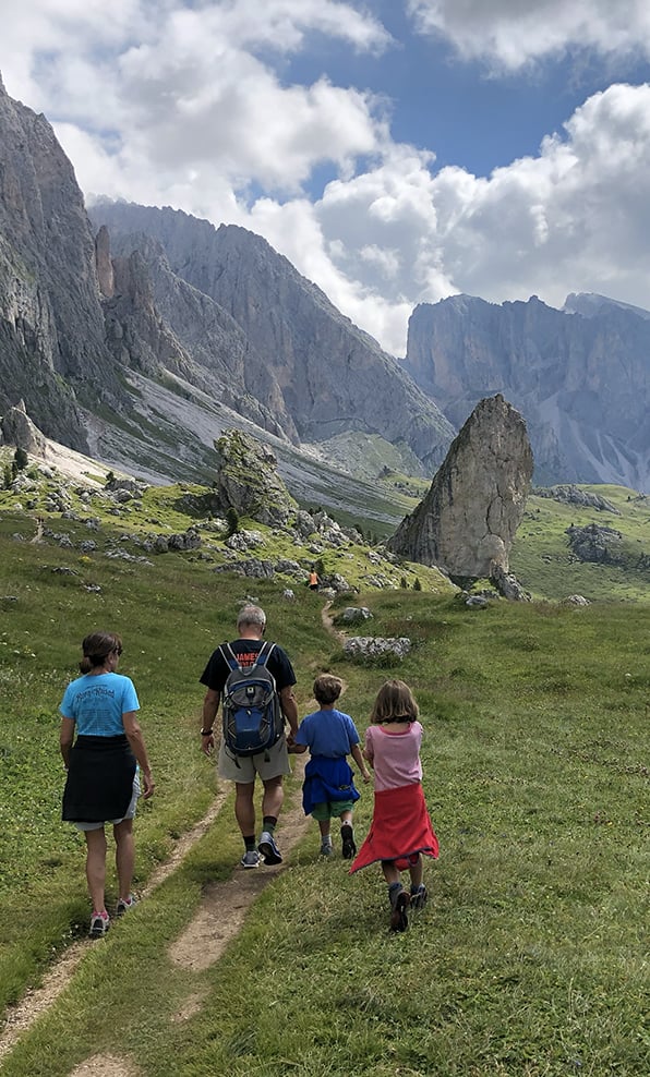 Quattro escursionisti camminano su sentiero erboso verso grandi rocce e picchi montani sotto cielo nuvoloso