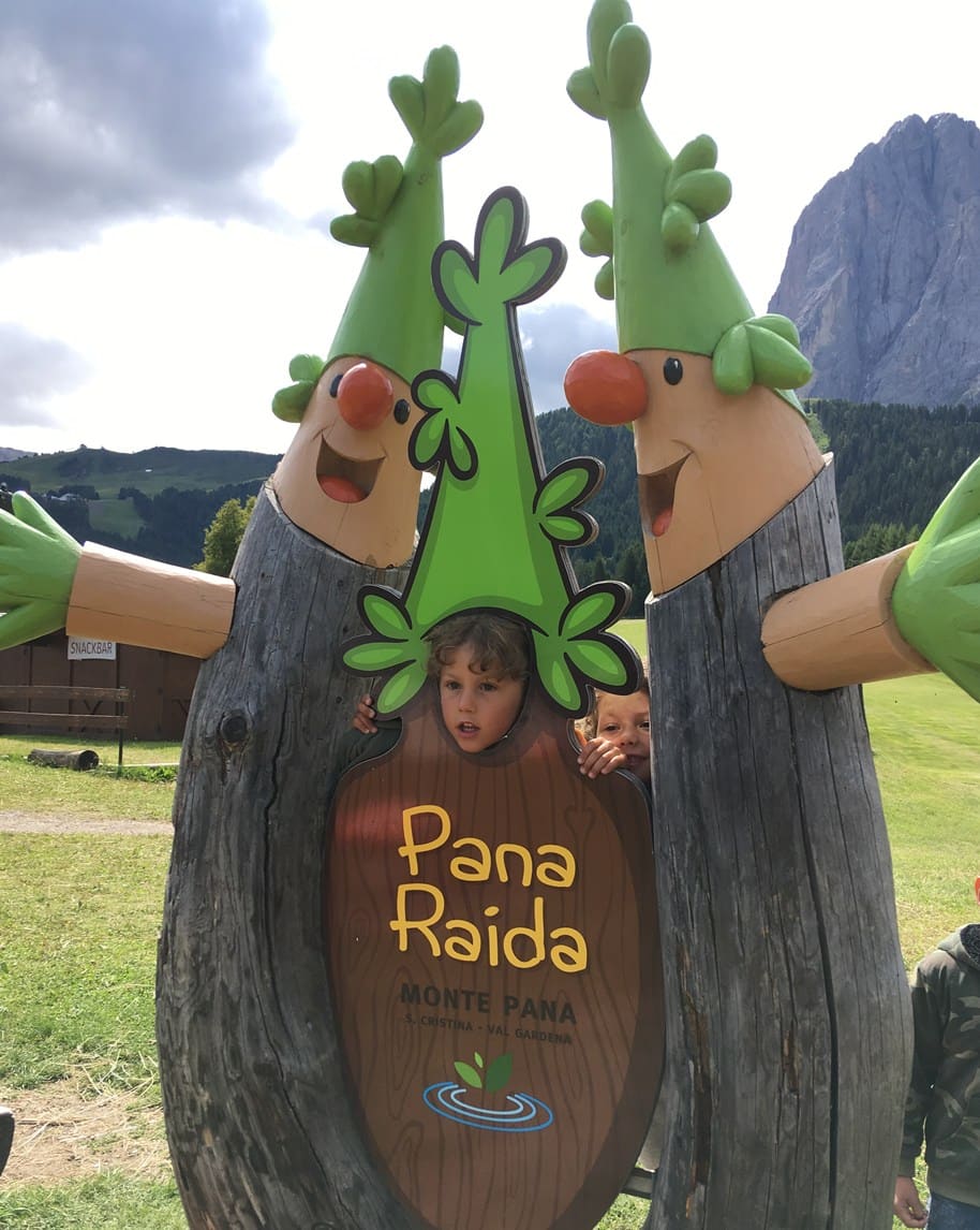 Bambini con la testa nelle sagome di legno a forma di alberi davanti al cartello 'Pana Raida'