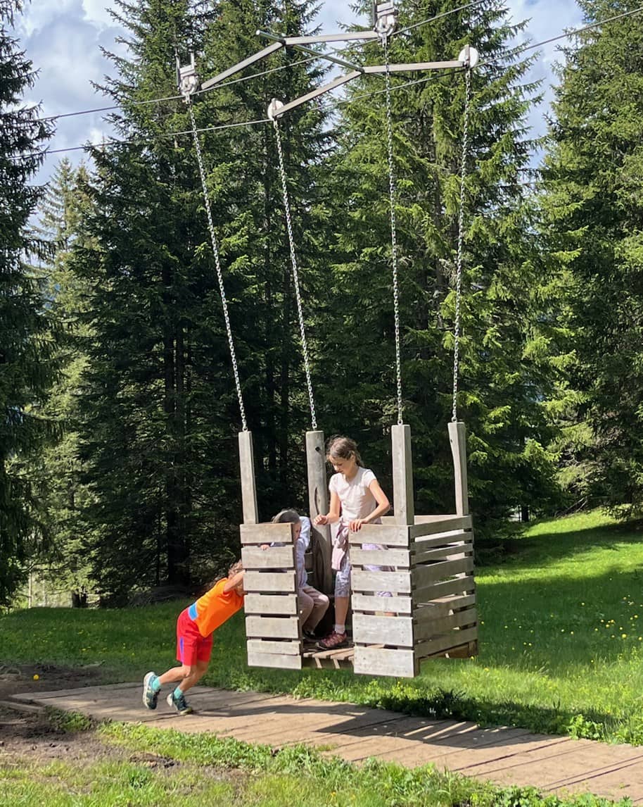 Tre bambini su un’altalena di legno sospesa; uno la spinge, bosco di conifere sullo sfondo