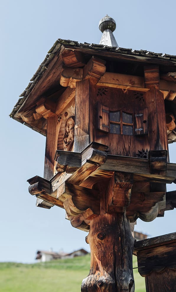 Casetta in legno intagliata su palo, tetto spiovente; prato e case alpine sfocate sullo sfondo
