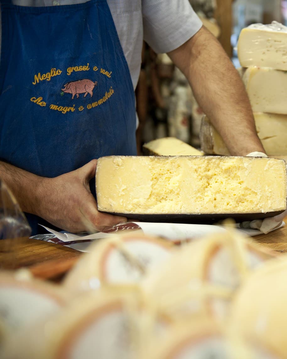 Addetto con grembiule che mostra una grande forma di formaggio tagliata, interno giallo friabile, forme sullo sfondo