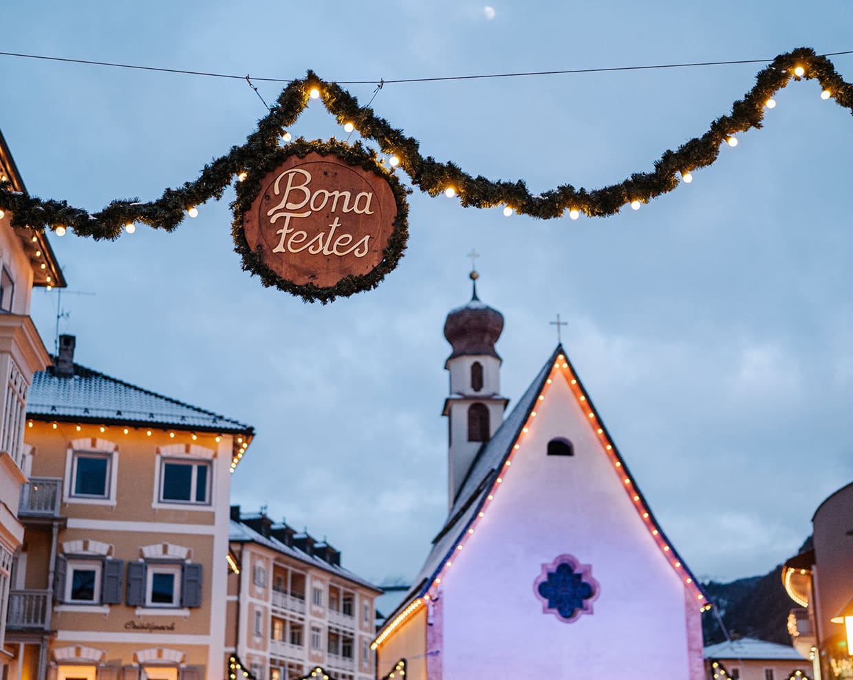 Borgo innevato con chiesa illuminata e ghirlanda sospesa con insegna 'Bona Festes' e luci natalizie