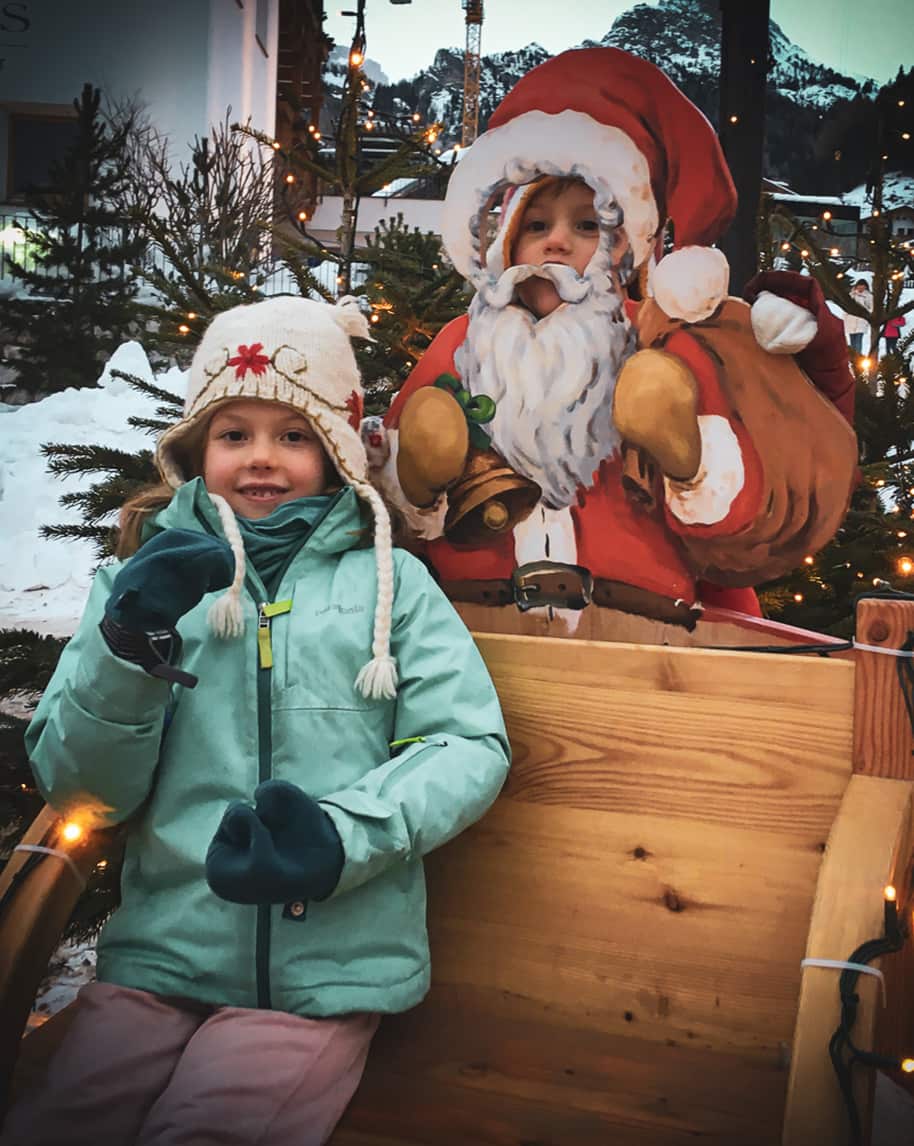 Bambina con giacca verde e cappello su slitta di legno accanto a sagoma di Babbo Natale con volto che spunta