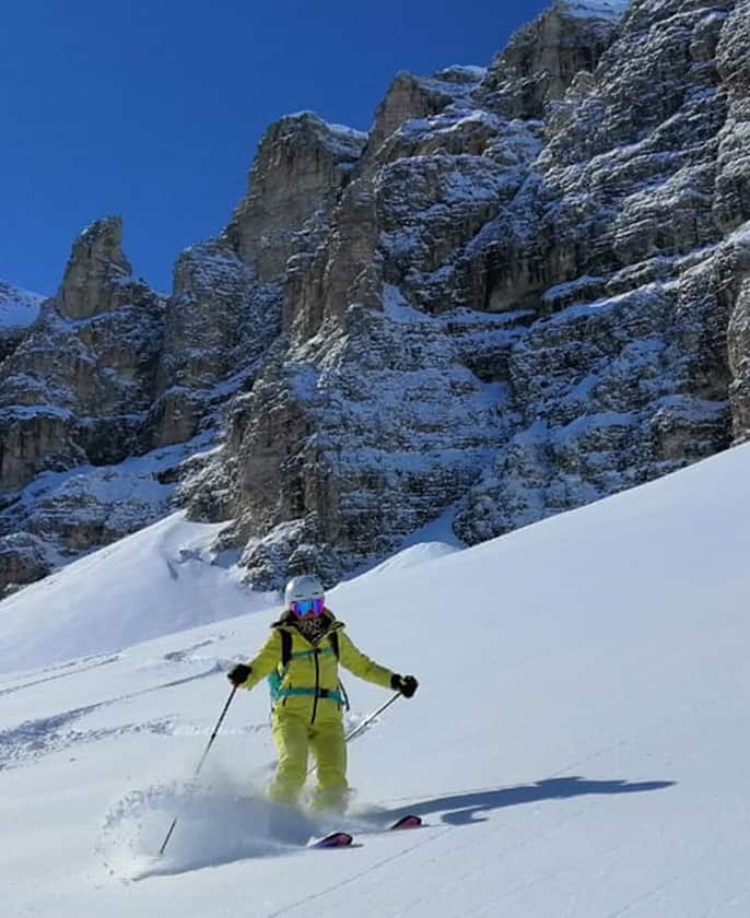 Sciatore in tuta gialla scende su neve fresca davanti a parete rocciosa innevata