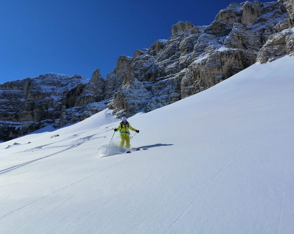 Sciatore in tuta gialla scende su neve fresca davanti a pareti rocciose innevate sotto cielo blu