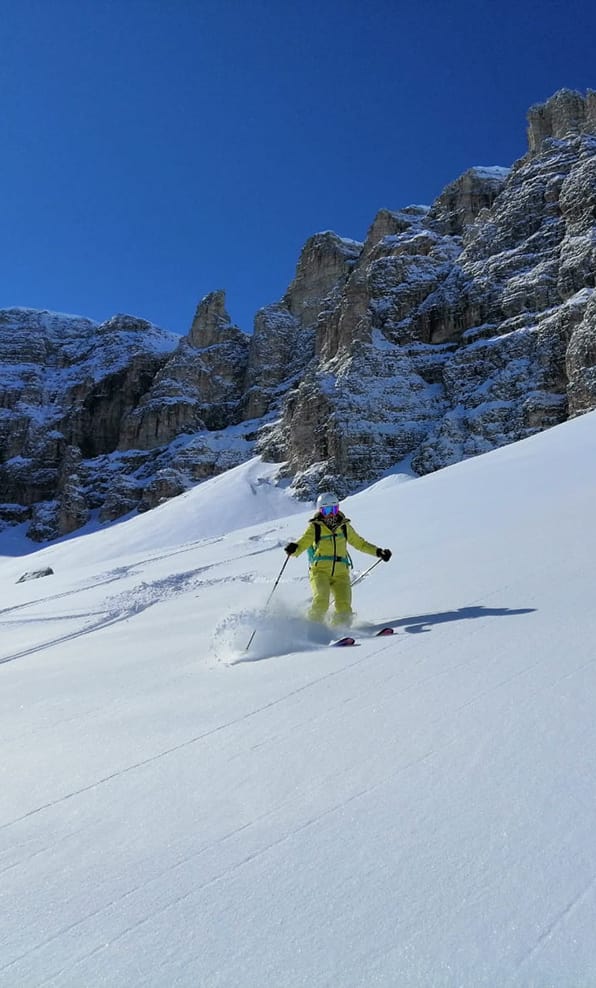 Sciatore in tuta gialla scende su neve fresca davanti a parete rocciosa innevata e cielo blu