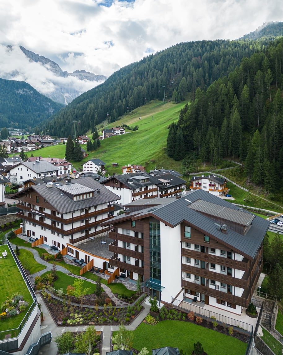 Hotel chalet a più piani in un paese alpino, prato e funivia su collina boscosa, montagne nuvolose sullo sfondo