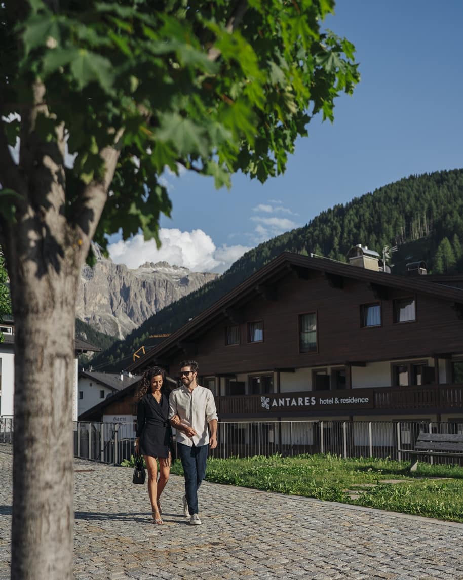Coppia che cammina braccio a braccio su un viale di ciottoli davanti all'Hotel ANTARES, montagne sullo sfondo