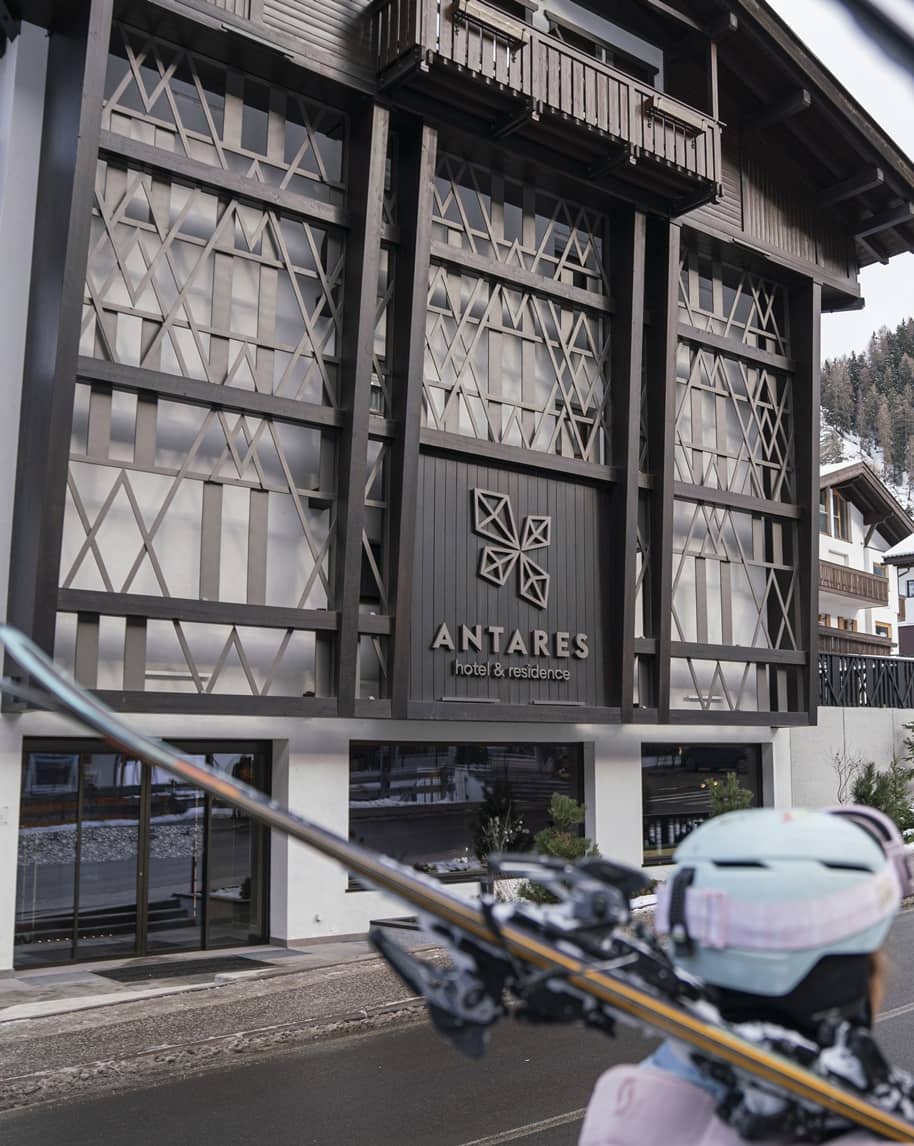 Edificio di Antares hotel & residence con facciata in legno; in primo piano persona con casco e sci sulla spalla