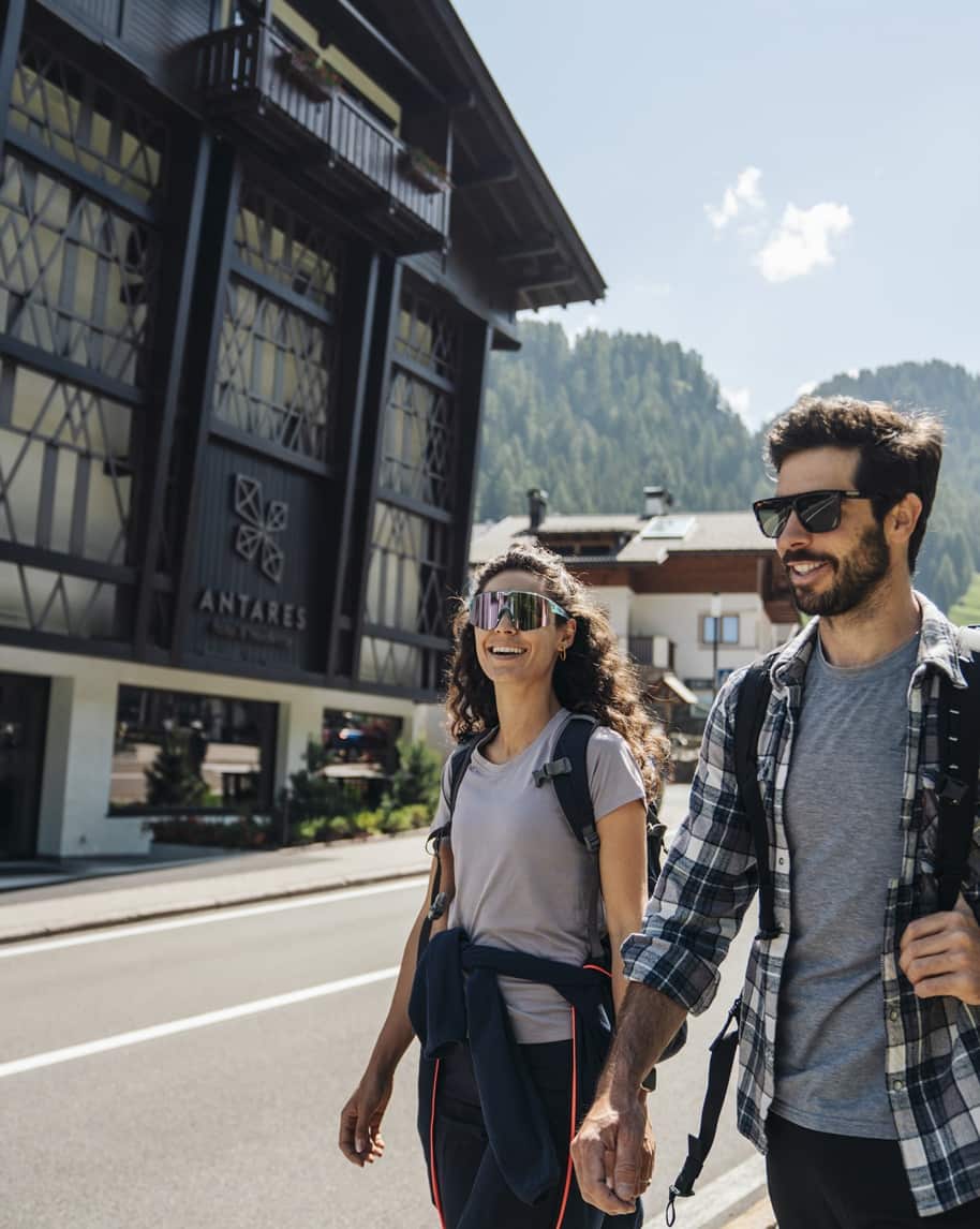 Due persone con zaini che camminano lungo una strada davanti all'Hotel Antares, montagne sullo sfondo