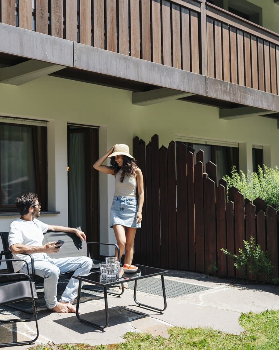 Coppia su terrazza: uomo seduto con telefono, donna con cappello in piedi vicino a tavolino con bicchieri e frutta