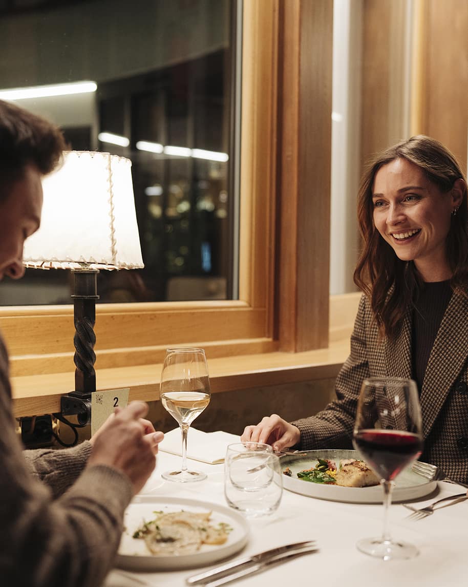 Coppia che cena al tavolo vicino alla finestra: donna sorridente, piatti e calici di vino
