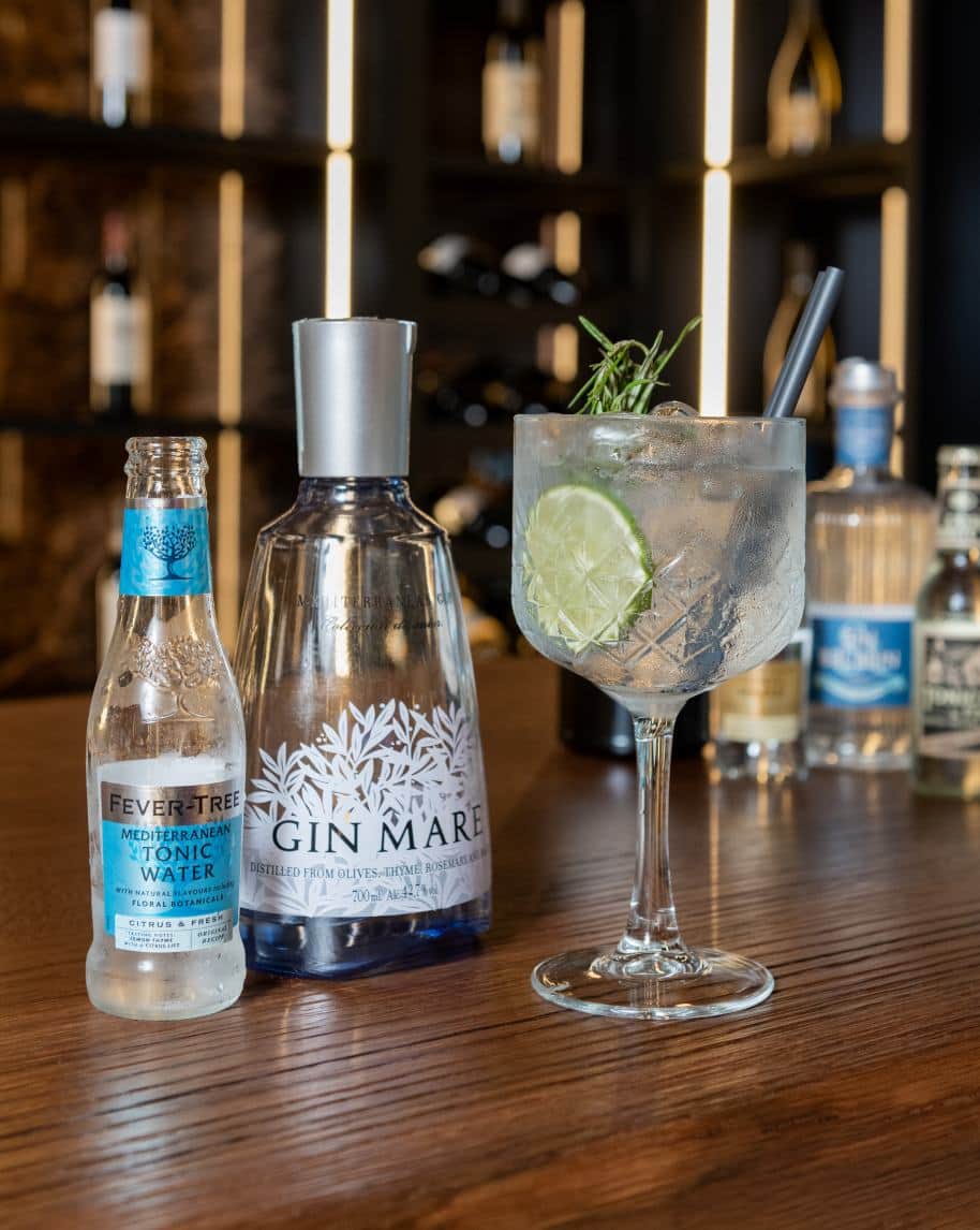 Bicchiere da gin con lime e rosmarino accanto a bottiglie di gin e tonica su bancone di legno