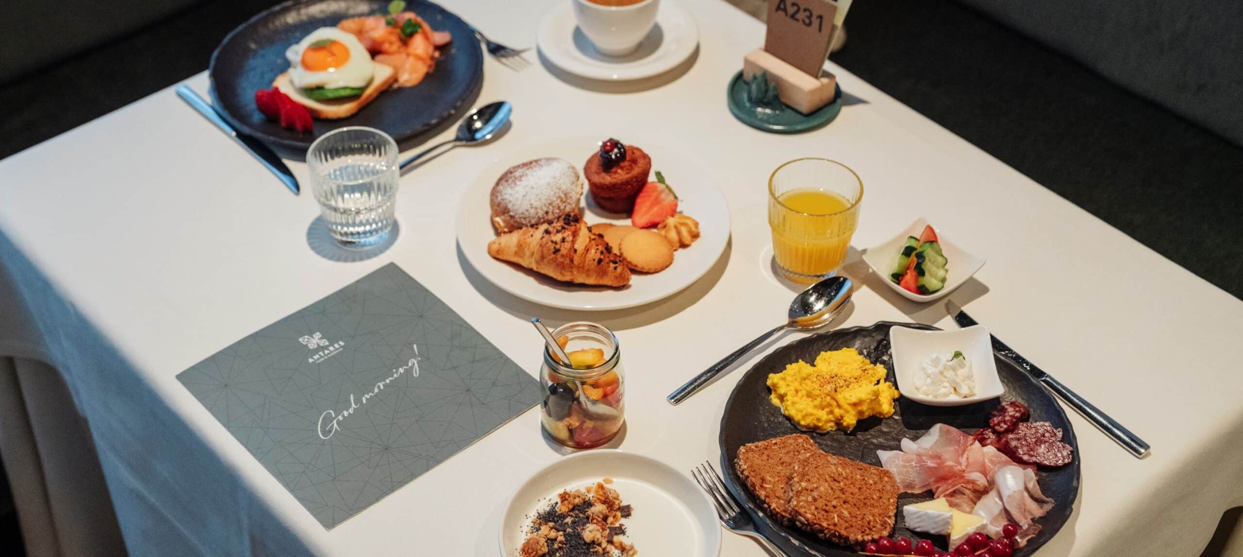 Tavola apparecchiata con piatti di colazione: pasticceria, croissant, uova strapazzate, salumi, succo e caffè