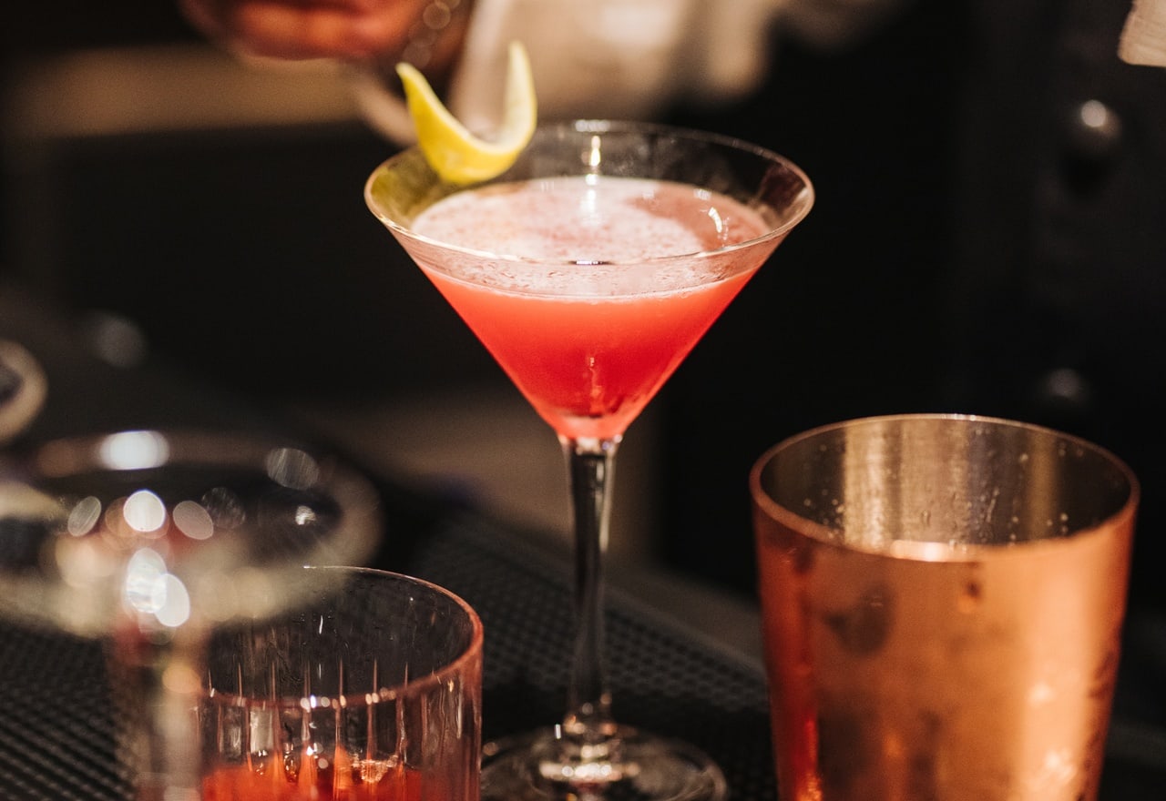 Calice a martini con cocktail rosa e scorza di limone a spirale, appoggiato su bancone accanto a bicchieri e shaker