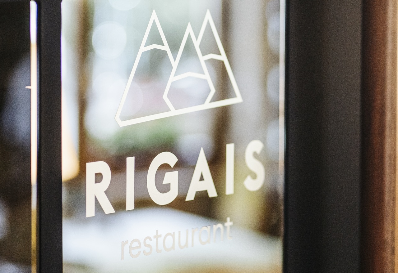 Vetro con logo a montagna e scritta RIGAIS restaurant, vista su interno sfocato