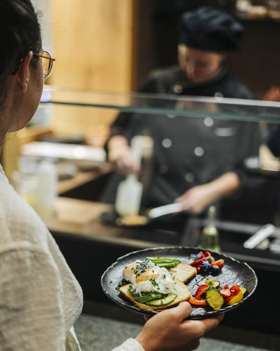 Persona tiene piatto di toast, uova in camicia, asparagi e verdure davanti al banco con cuoco