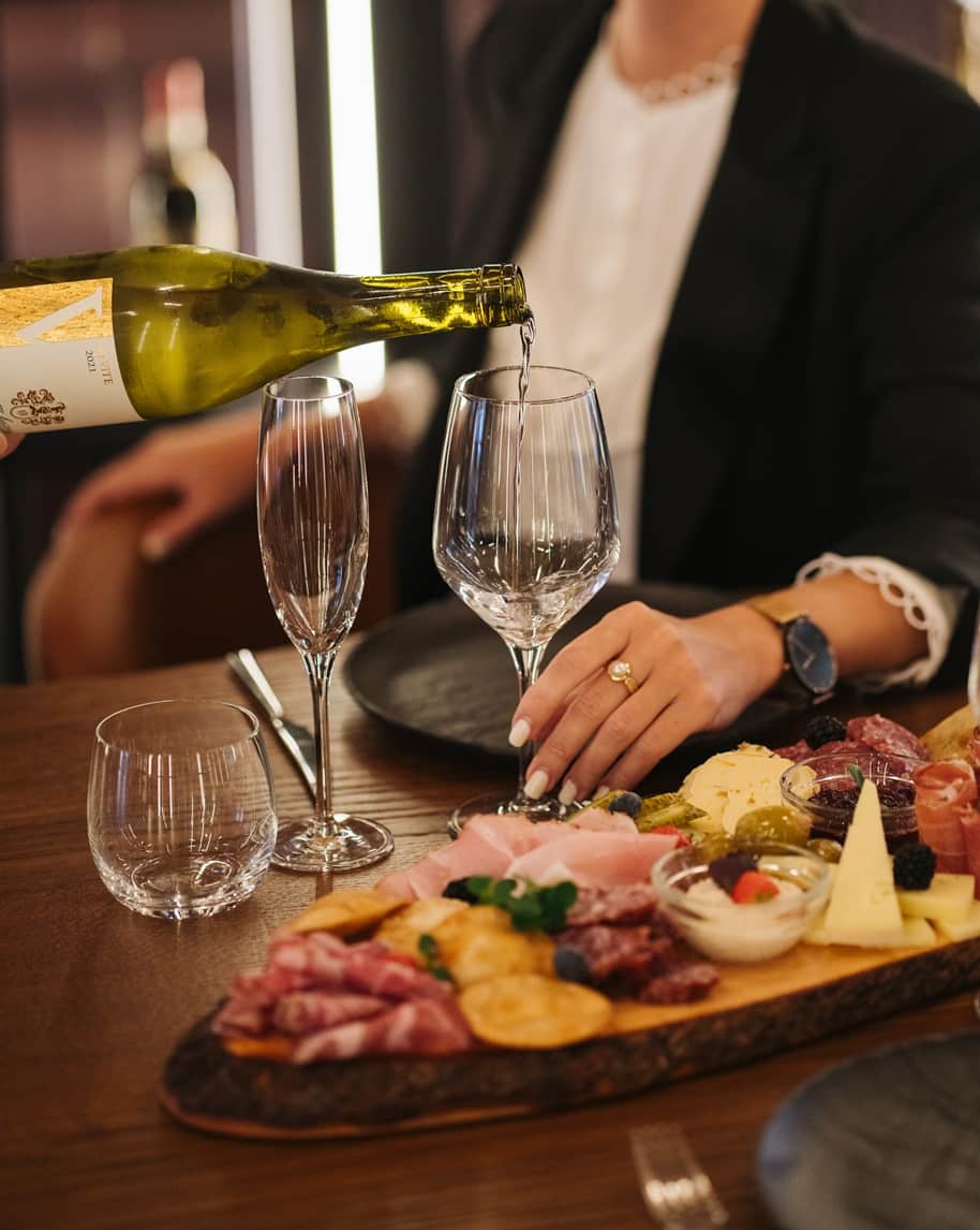 Bottiglia che versa vino in un calice; mano con anello vicino a un tagliere di salumi e formaggi
