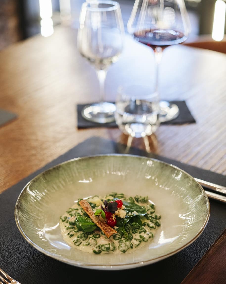 Piatto con gnocchi verdi in crema, foglie e bacche rosse, su tavolo con calici di vino sullo sfondo