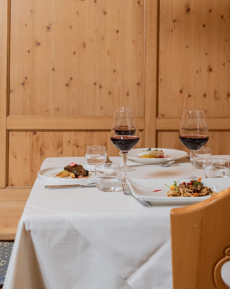 Tavolo apparecchiato con tovaglia bianca, piatti gourmet e due calici di vino rosso davanti a parete in legno