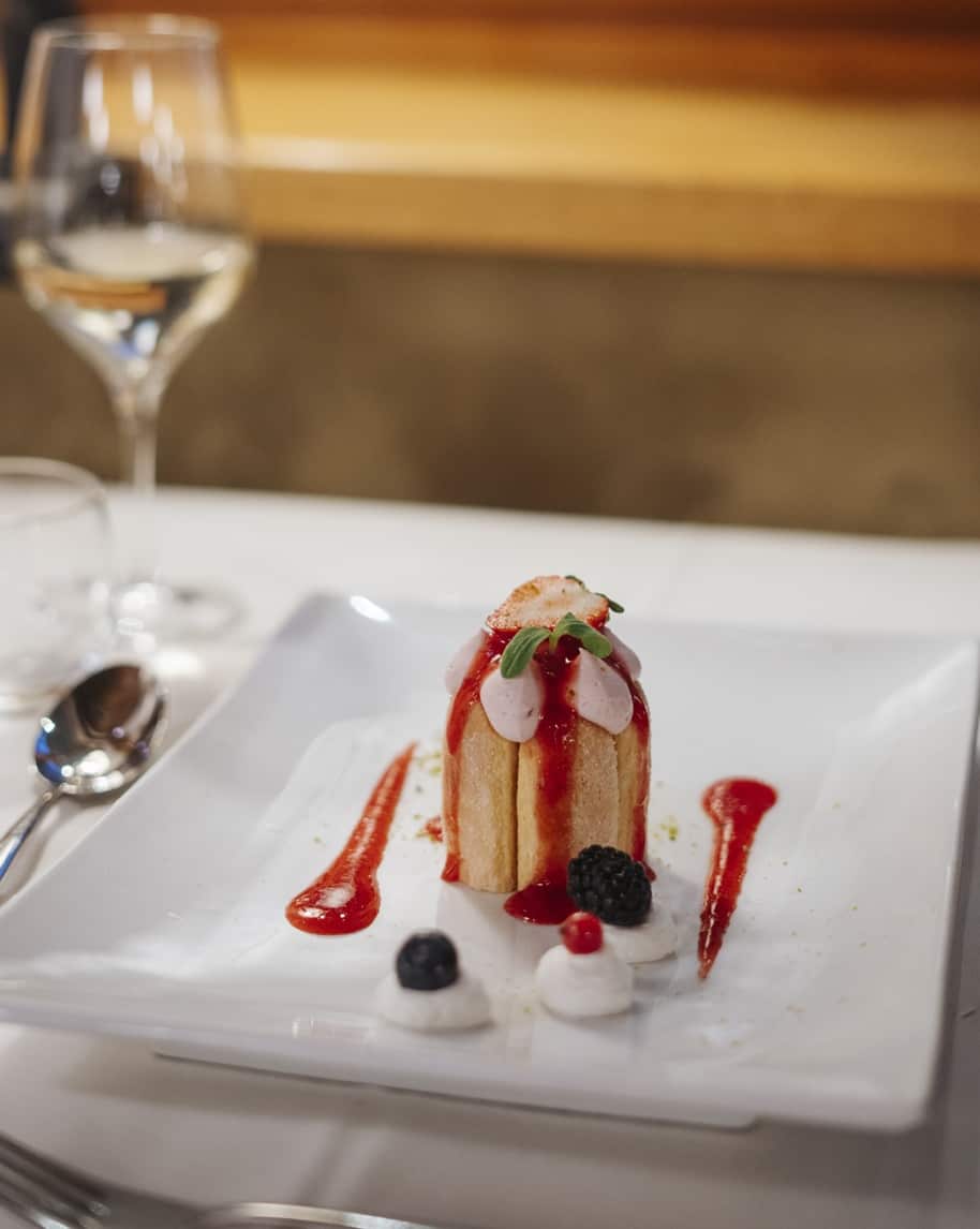 Dolce a cilindro con crema rosa e salsa di frutti rossi su piatto bianco, bicchiere di vino sullo sfondo