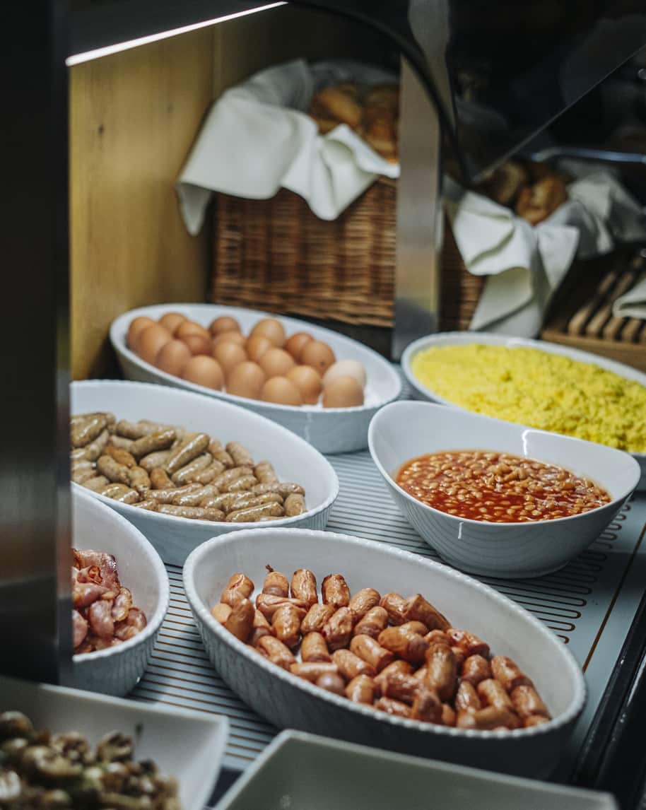 Buffet colazione con ciotole di salsicce, uova intere, fagioli al pomodoro, uova strapazzate e cestino di panini