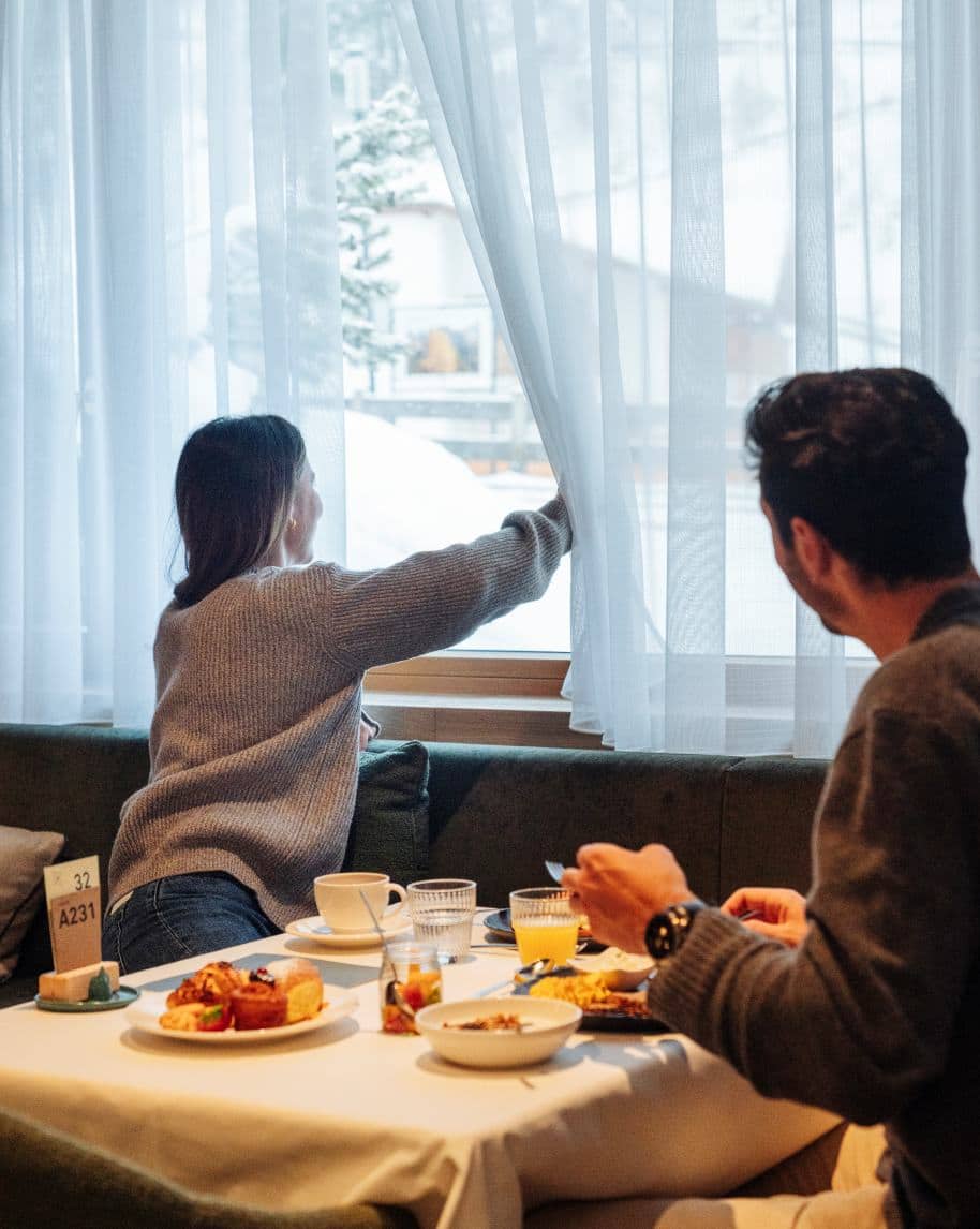 Donna tira la tenda per guardare fuori mentre un uomo fa colazione a un tavolo; neve visibile fuori