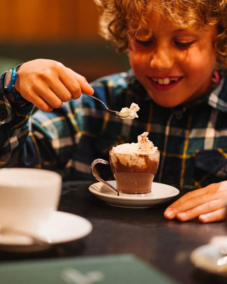 Bambino raccoglie panna con un cucchiaio da una tazza di cioccolata calda su piattino