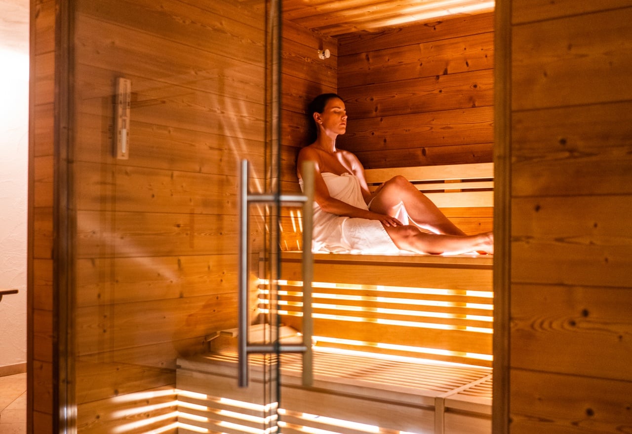 Donna seduta su panca in sauna di legno, avvolta in asciugamano, occhi chiusi e luce calda