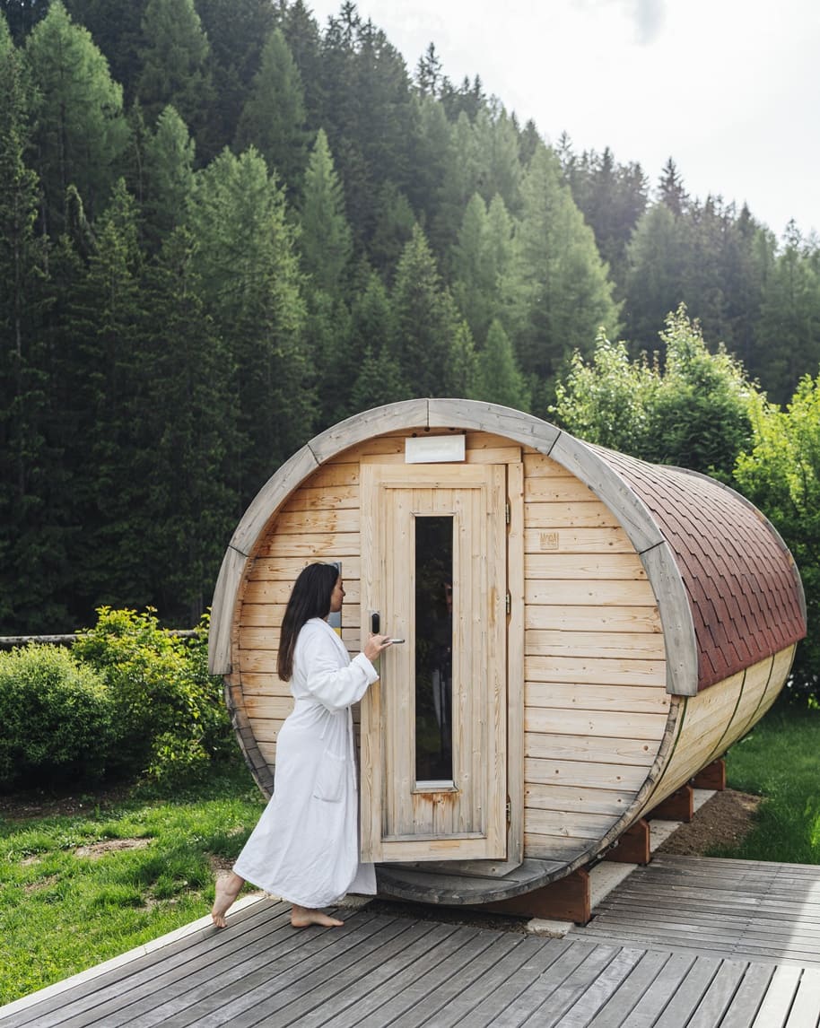 Donna in accappatoio apre porta di sauna cilindrica in legno su deck, boschi sullo sfondo