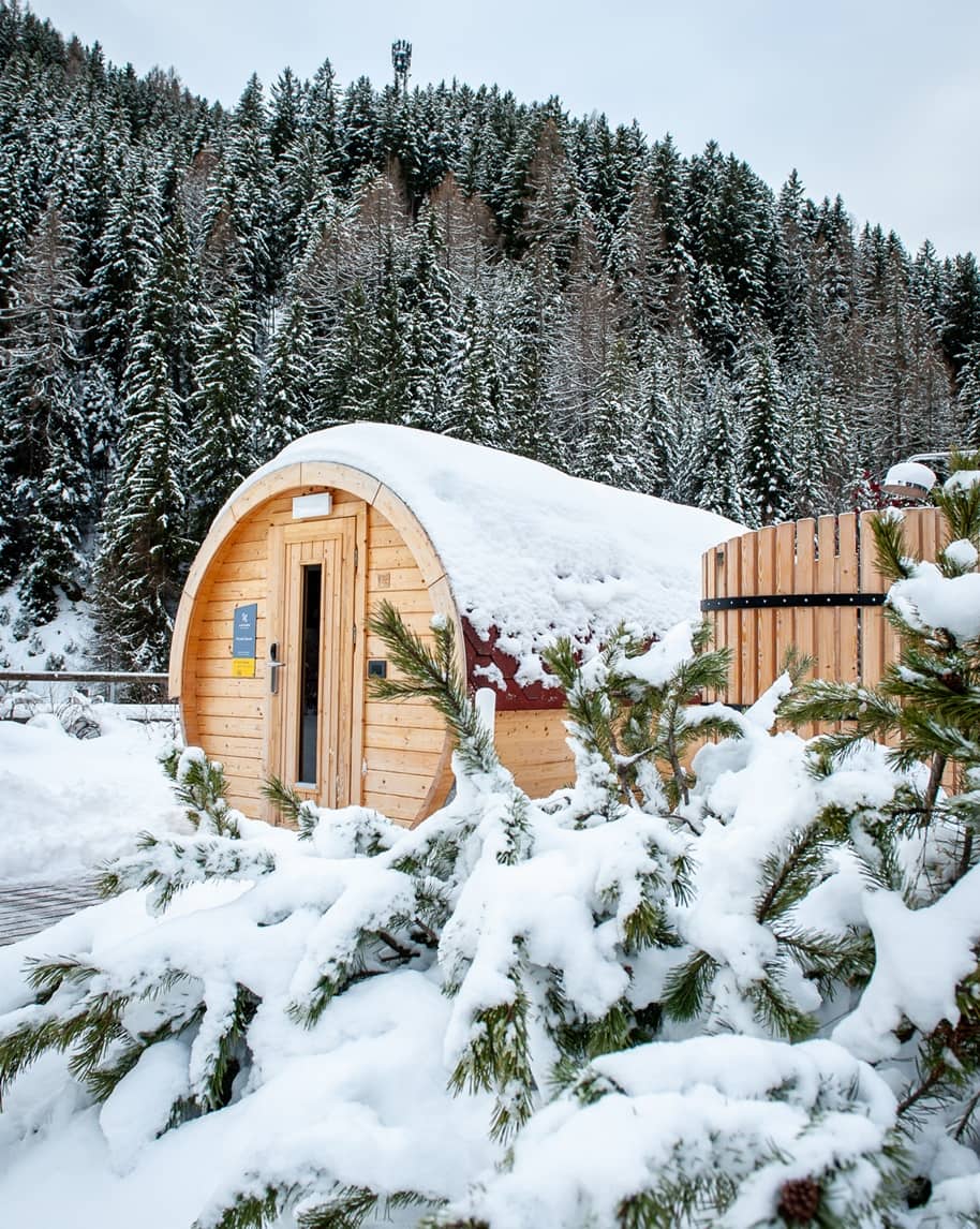 Struttura in legno a forma di botte coperta di neve, pini innevati in primo piano e foresta sullo sfondo