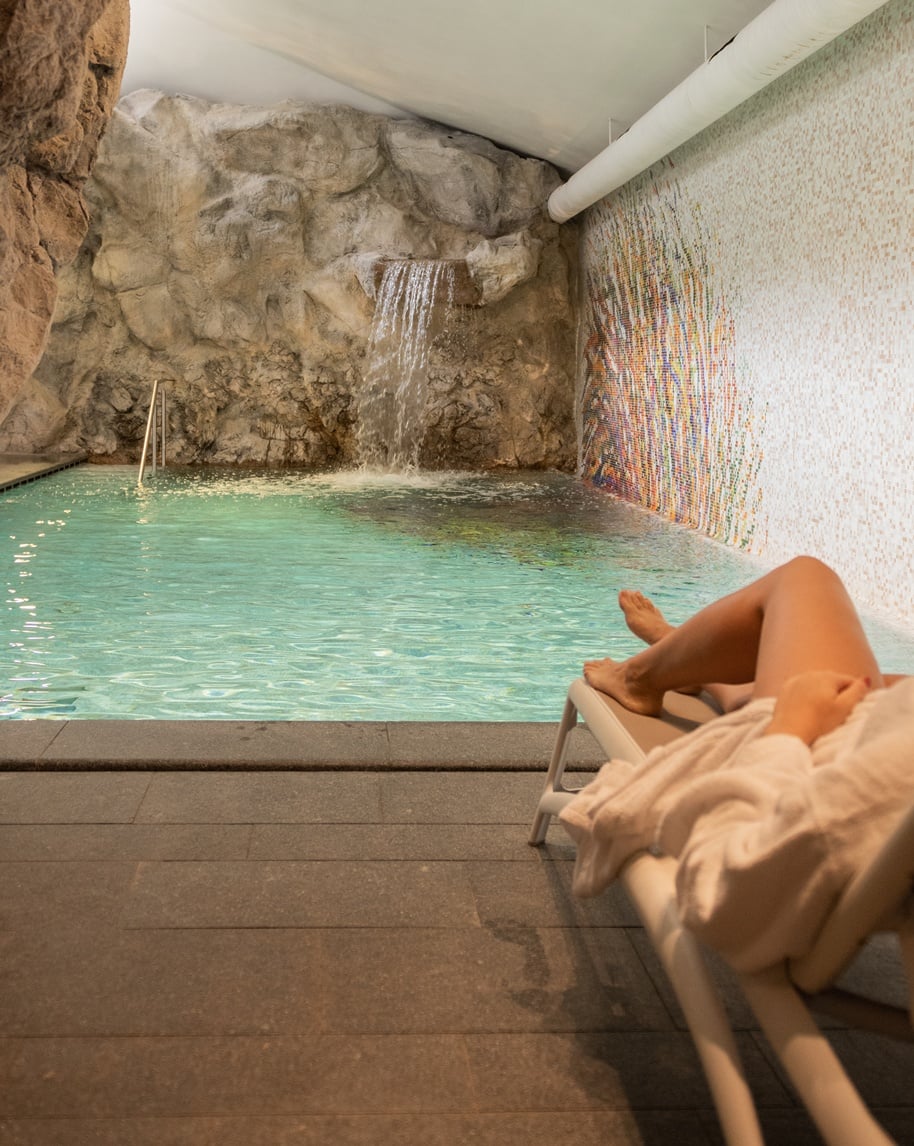 Piscina interna con cascata su parete rocciosa, mosaico laterale e persona distesa su lettino in accappatoio
