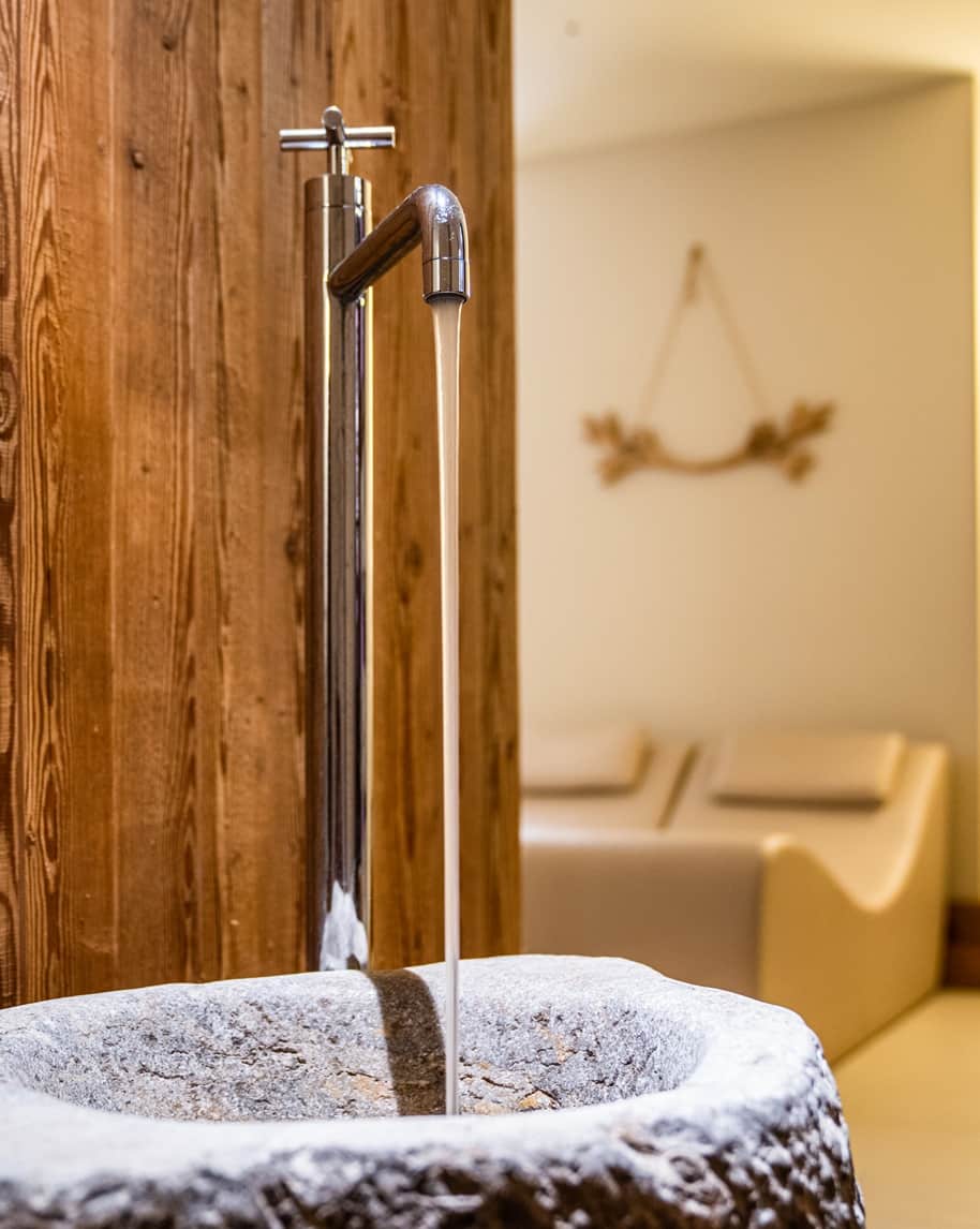 Rubinetto alto inox che lascia scorrere acqua in una vasca di pietra davanti a parete in legno e divanetti sfocati
