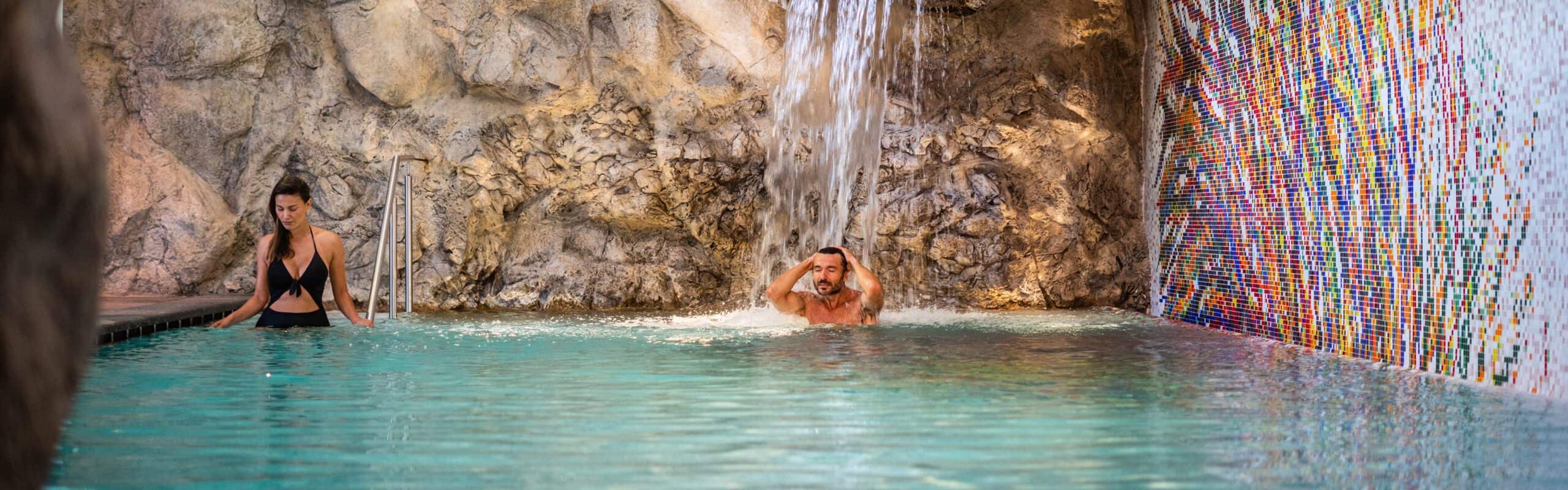 Uomo sotto cascata in piscina interna con parete rocciosa, donna accanto a scaletta e mosaico colorato