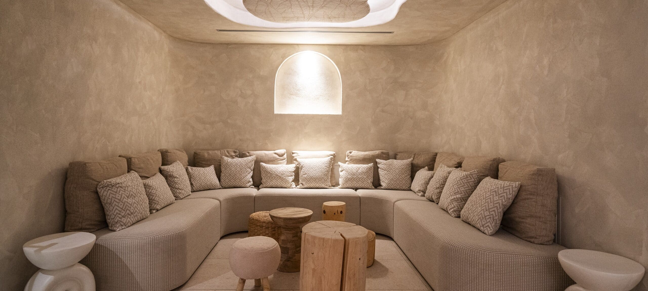 Salotto con divano curvo angolare, cuscini multipli, sgabelli e tavolini in legno e nicchia ad arco illuminata