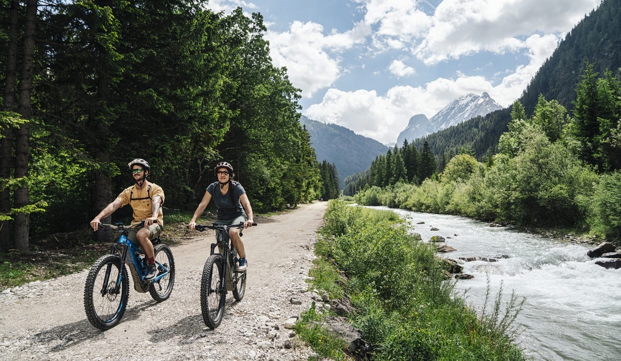 Due persone in bici su strada sterrata lungo un fiume, foresta a sinistra e montagne innevate sullo sfondo