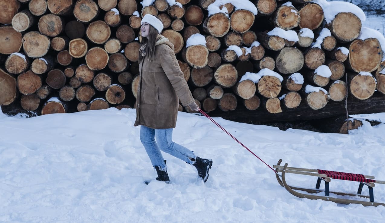 Donna con berretto bianco trascina una slitta di legno sulla neve davanti a una pila di tronchi