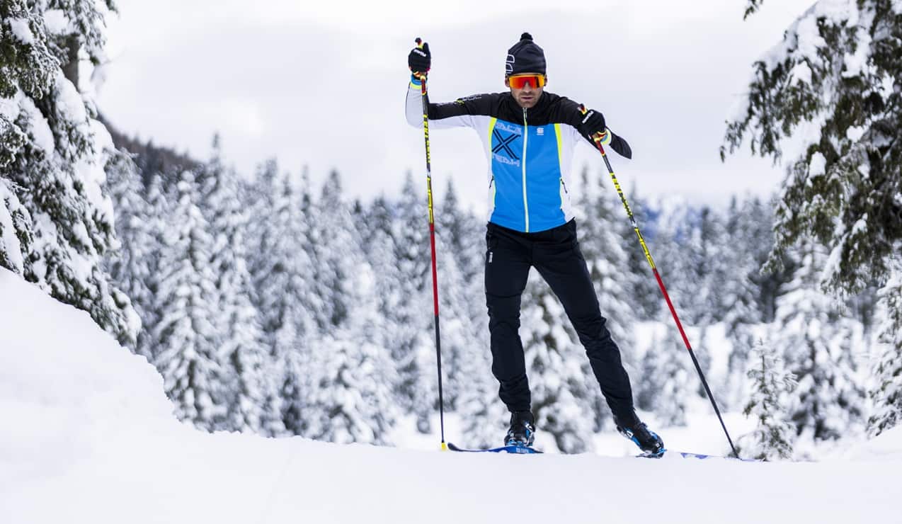 Sciatore con giacca blu scia su pista di fondo tra alberi innevati