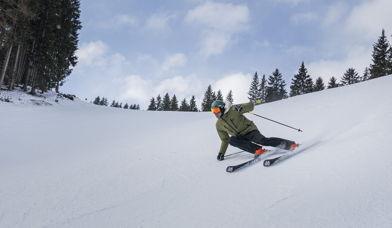 Sciatore in curva su pista innevata, alberi sullo sfondo e cielo nuvoloso
