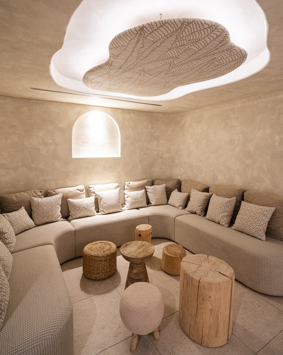 Salotto semicircolare con divano beige e cuscini, tavolini e sgabelli in legno, soffitto con illuminazione decorativa