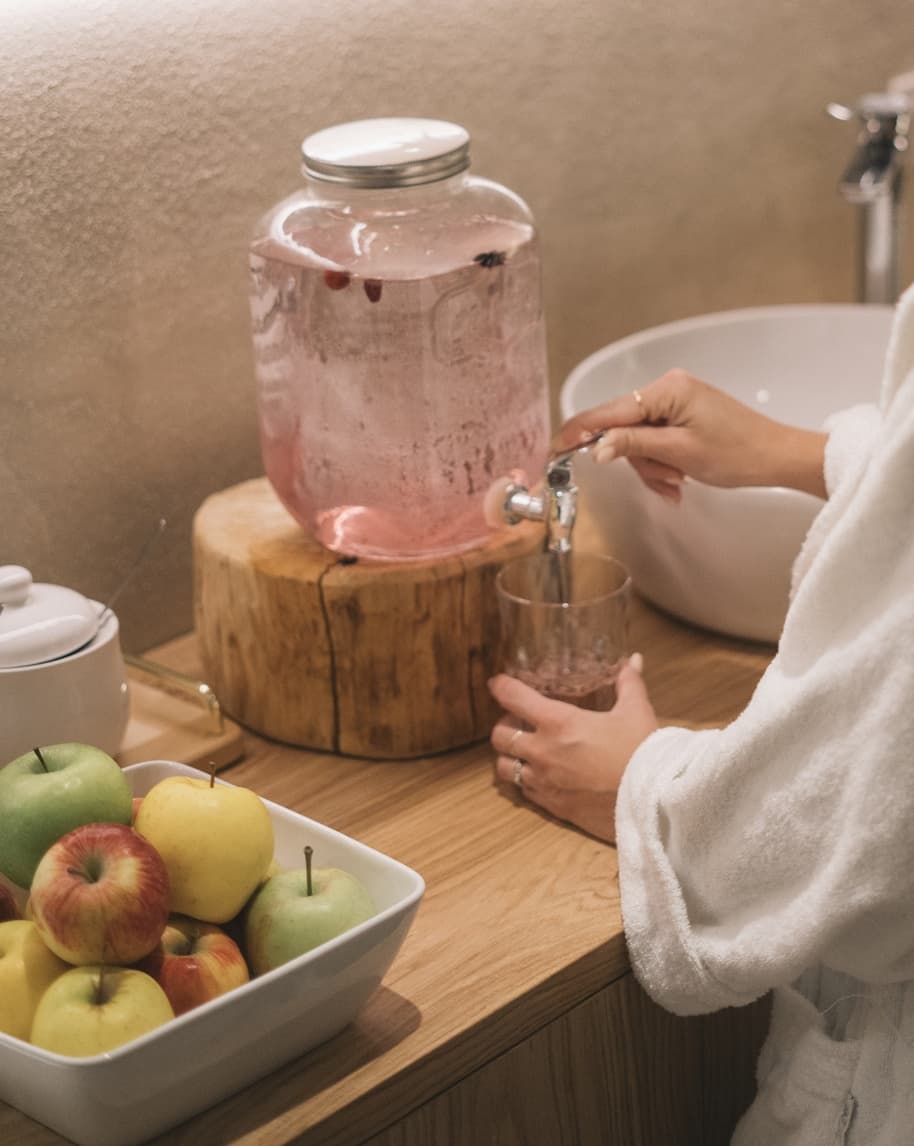 Dispenser in vetro con bevanda rosa, persona in accappatoio versa in un bicchiere su piano di legno, ciotola di mele
